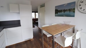 Una cocina moderna con armarios blancos, una mesa de comedor de madera y sillas blancas. Un reloj grande está colgado en la pared.