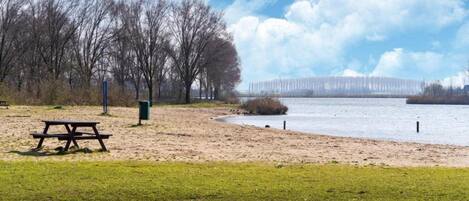 Un parc avec une table de pique-nique et un banc, une zone sablonneuse et une étendue d’eau avec un pont en arrière-plan.