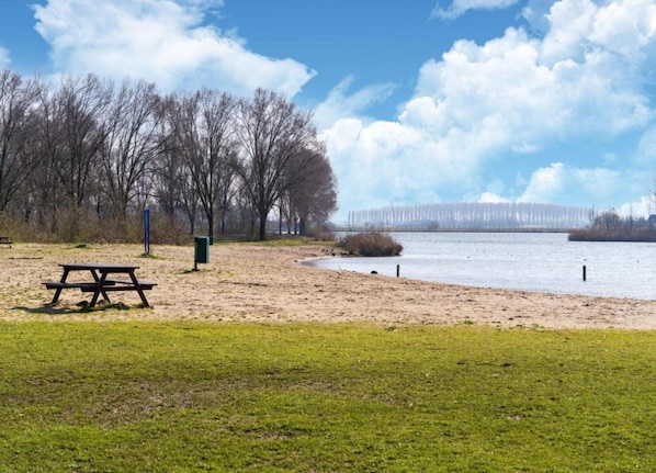 Een park met een picknicktafel en een bankje, een zandig gebied en een waterpartij met een brug op de achtergrond.