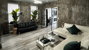 A modern living room with a black sofa, a white sofa, and a glass coffee table. There are two potted plants and a radiator.