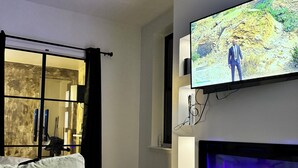 A modern living room with a flat-screen TV mounted on the wall, a fireplace with a blue light, a sofa, and a view through a glass door to an outdoor area.