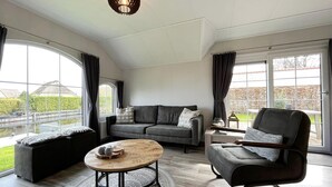 Un salon moderne avec un canapé gris, une table basse ronde et un fauteuil noir. De grandes fenêtres offrent une vue sur l’extérieur.