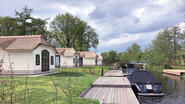 Une propriété située au bord d’un canal, avec une rangée de bâtiments blancs, une jetée en bois et un bateau amarré sur l’eau.