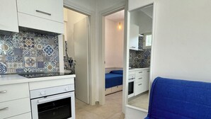 A compact kitchen with white cabinets and a patterned backsplash, a small oven, and a blue sofa in a narrow room with a door leading to another area. - Miramare House - Trilo 5 D (Puglia)
