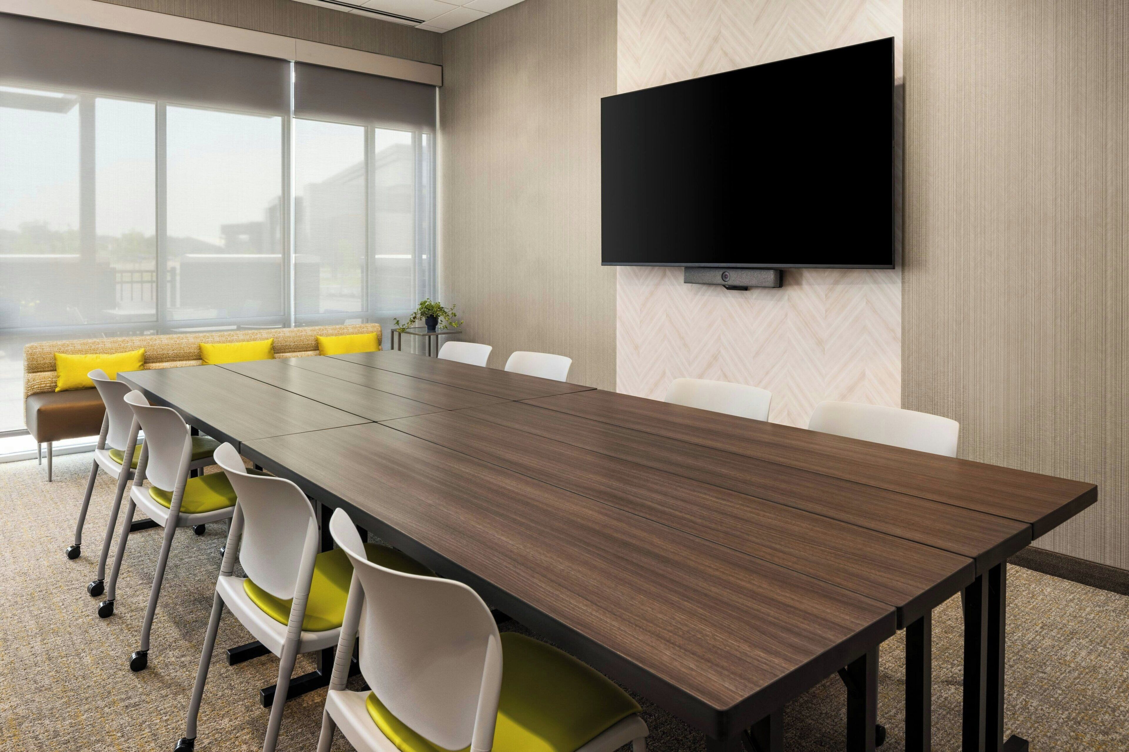 A conference room with a long table, white chairs with yellow cushions, a sofa with yellow cushions, and a large TV on the wall.
