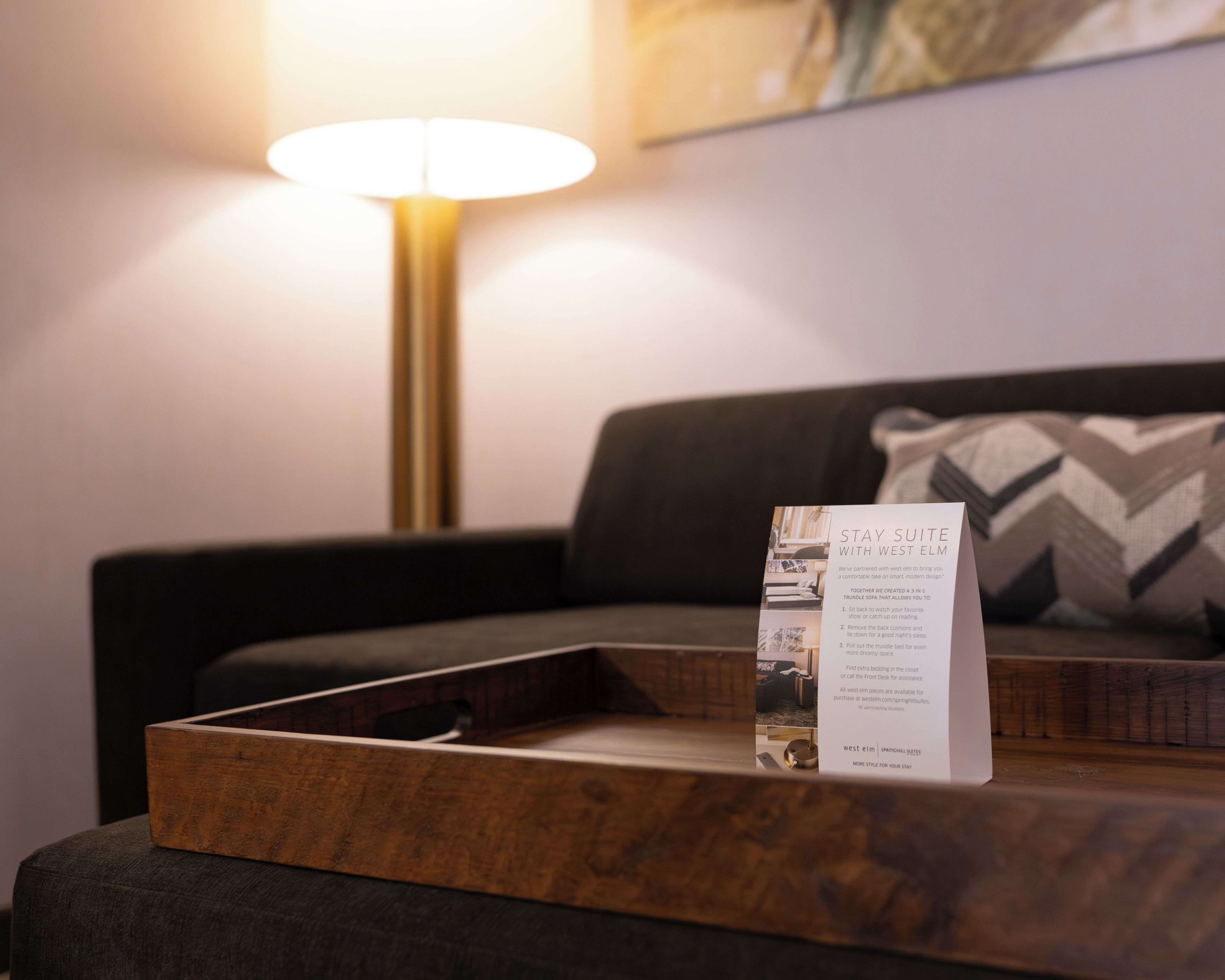 Una bandeja de madera sobre un otoman oscuro con un letrero sobre Stay Suite con West Elm.