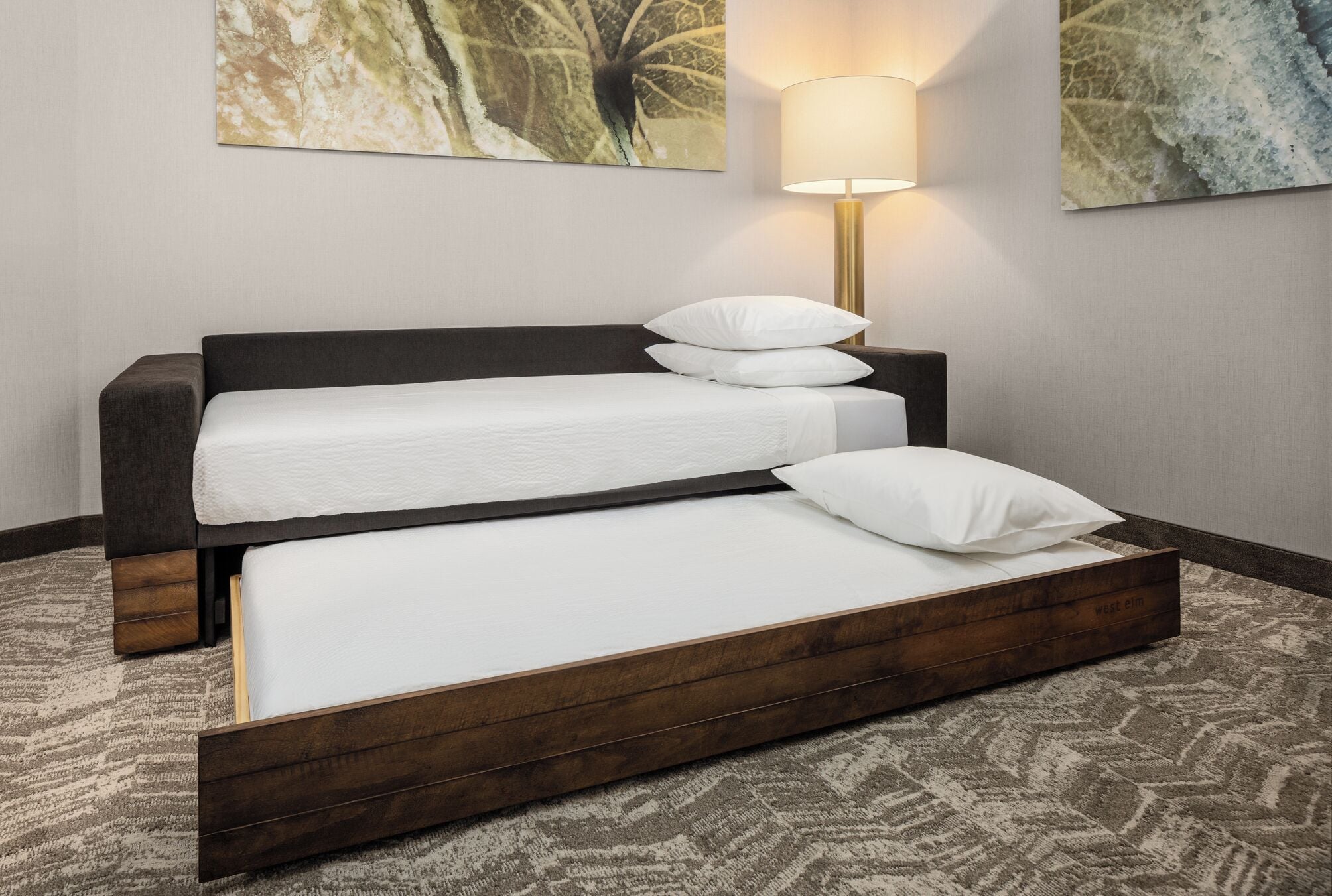 A double bed with a wooden frame, white bedding, and a headboard. There is a wall-mounted lamp and two abstract paintings above the bed.