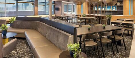 Un hall d’hôtel moderne doté d’un grand espace salon, de plusieurs tables à manger et d’une grande fenêtre offrant une vue sur l’extérieur.