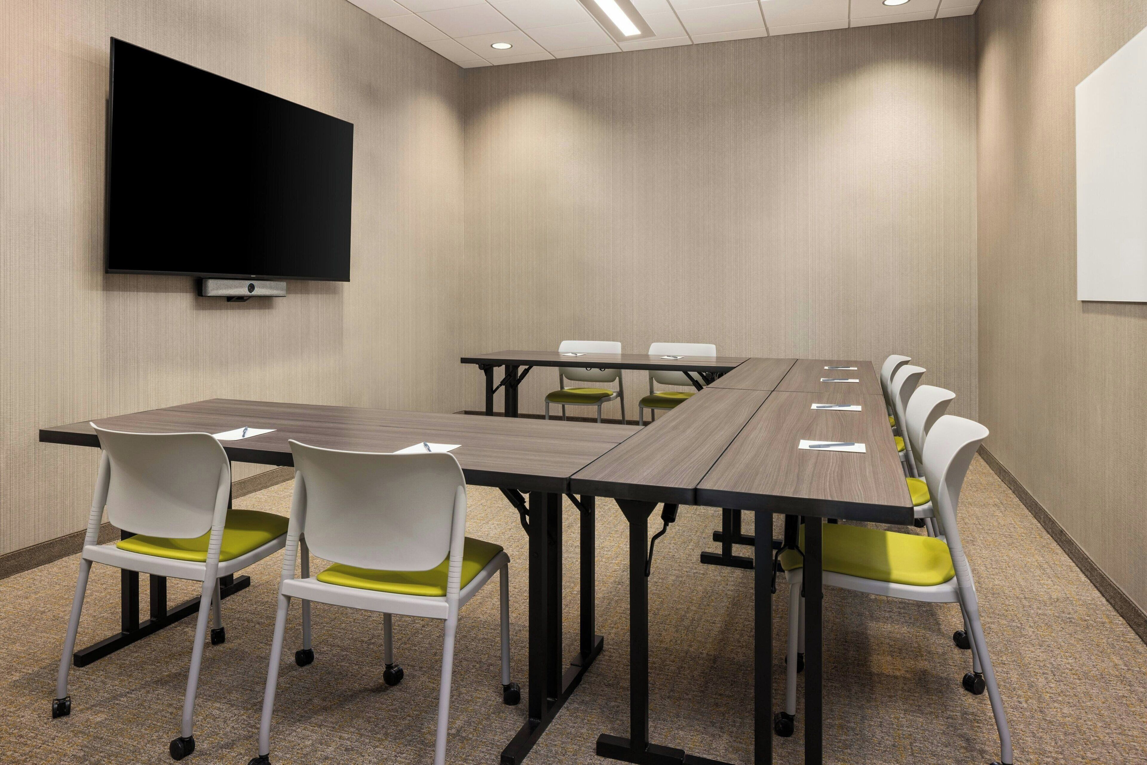 A conference room with a long table, white chairs with yellow cushions, a flat-screen TV on the wall, and a whiteboard.