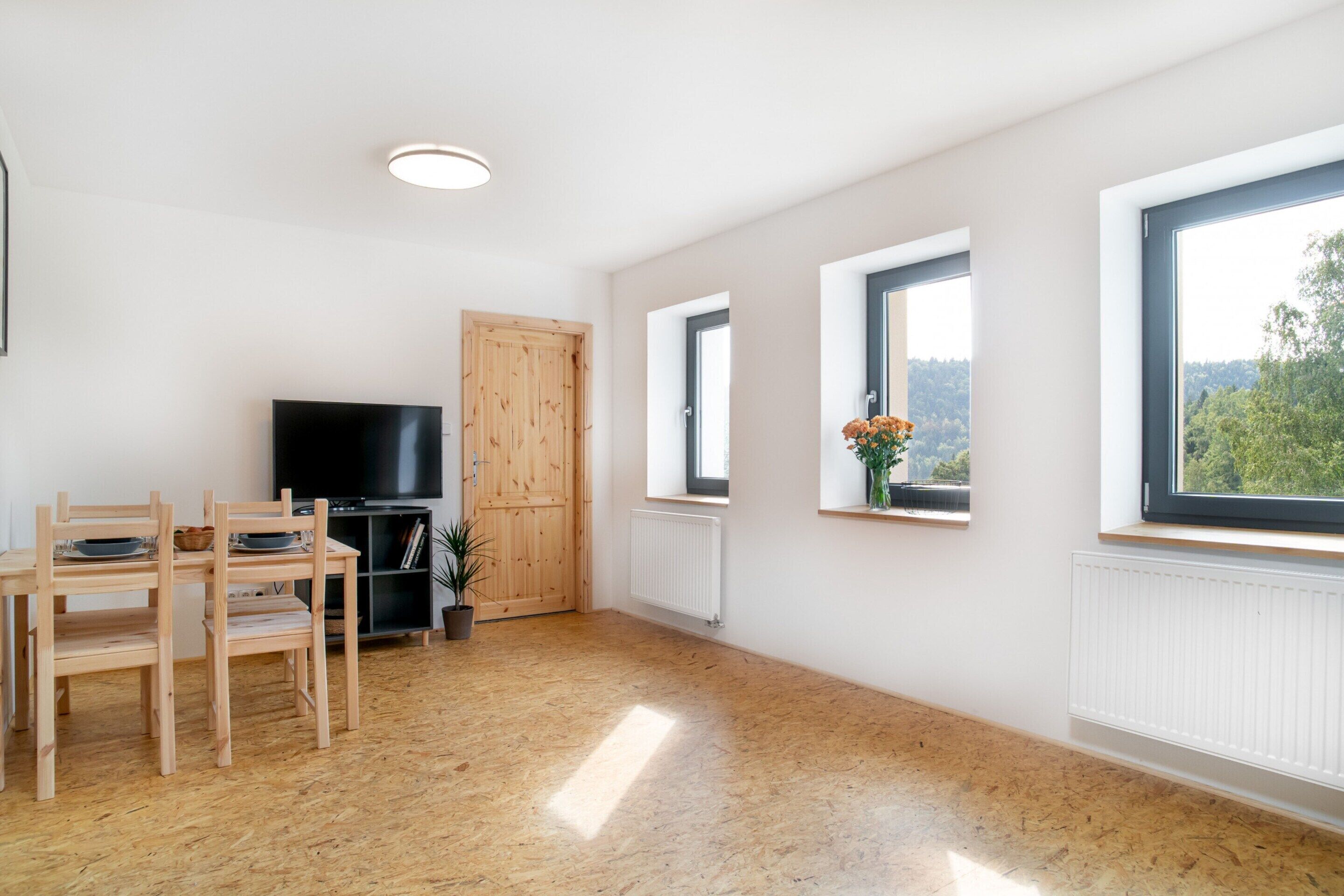 A spacious room with a wooden floor, a TV, a wooden door, and windows with views of greenery.