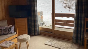A cozy room with a wooden interior, a television on a wooden cabinet, a sofa with a cushion, a small stool, and a window with a view of a snowy landscape.