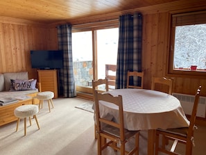A cozy room with a wooden interior, a television, a sofa with a dog-themed pillow, a dining table with chairs, and a window with a view of snow-covered trees.