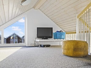 A room with a sloped ceiling, a television on a stand, a blue storage bin, and a patterned ottoman. - 5 Star Holiday Home in Lokken (Lokken)