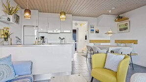 A modern kitchen with white cabinets, a central island, and pendant lighting. There is a dining area with a yellow chair and a table, and a living area with a sofa and a coffee table. - 5 Star Holiday Home in Lokken (Lokken)