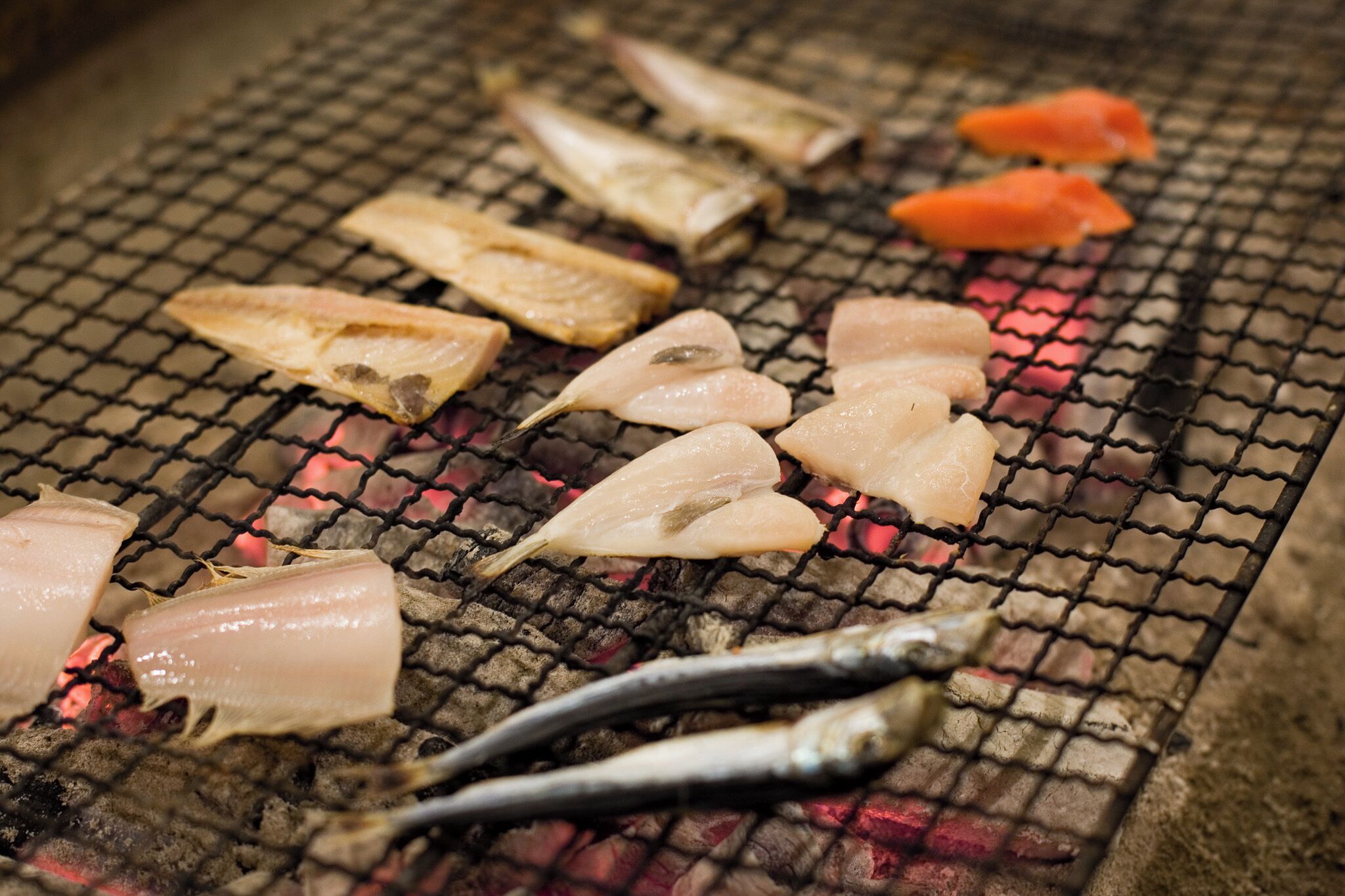 Various fish being grilled on a metal grate over an open flame.