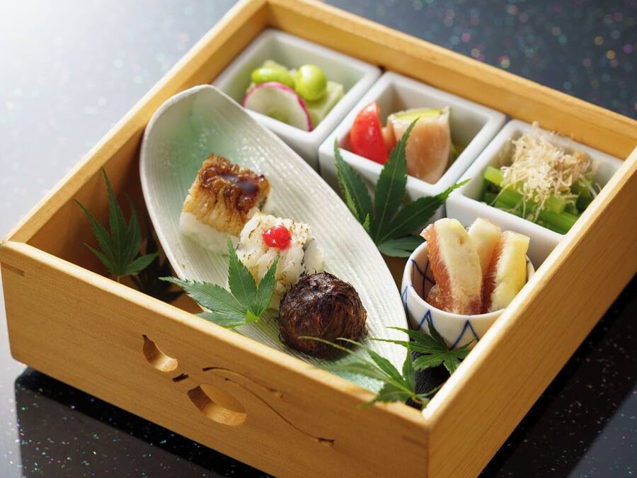 A wooden box containing various food items, including sushi, pickled vegetables, and a bowl of rice.