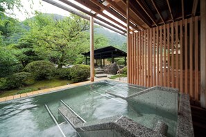 Ein traditionelles japanisches Onsen mit Holzbau, einem Steinbecken für Thermalwasser und Blick auf üppige Vegetation und Berge.