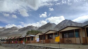 A row of wooden cabins with large windows, set against a backdrop of mountains and a partly cloudy sky. - Ecstasy Huts Pangong (Leh)