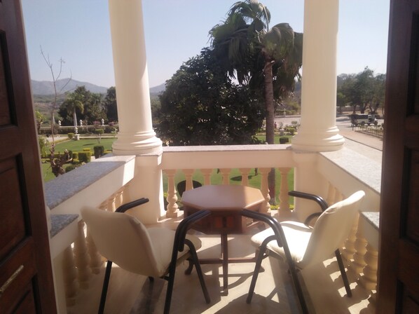 A balcony with a table and chairs, overlooking a landscaped garden and palm trees. - Balaram Palace Resort (Palanpur)