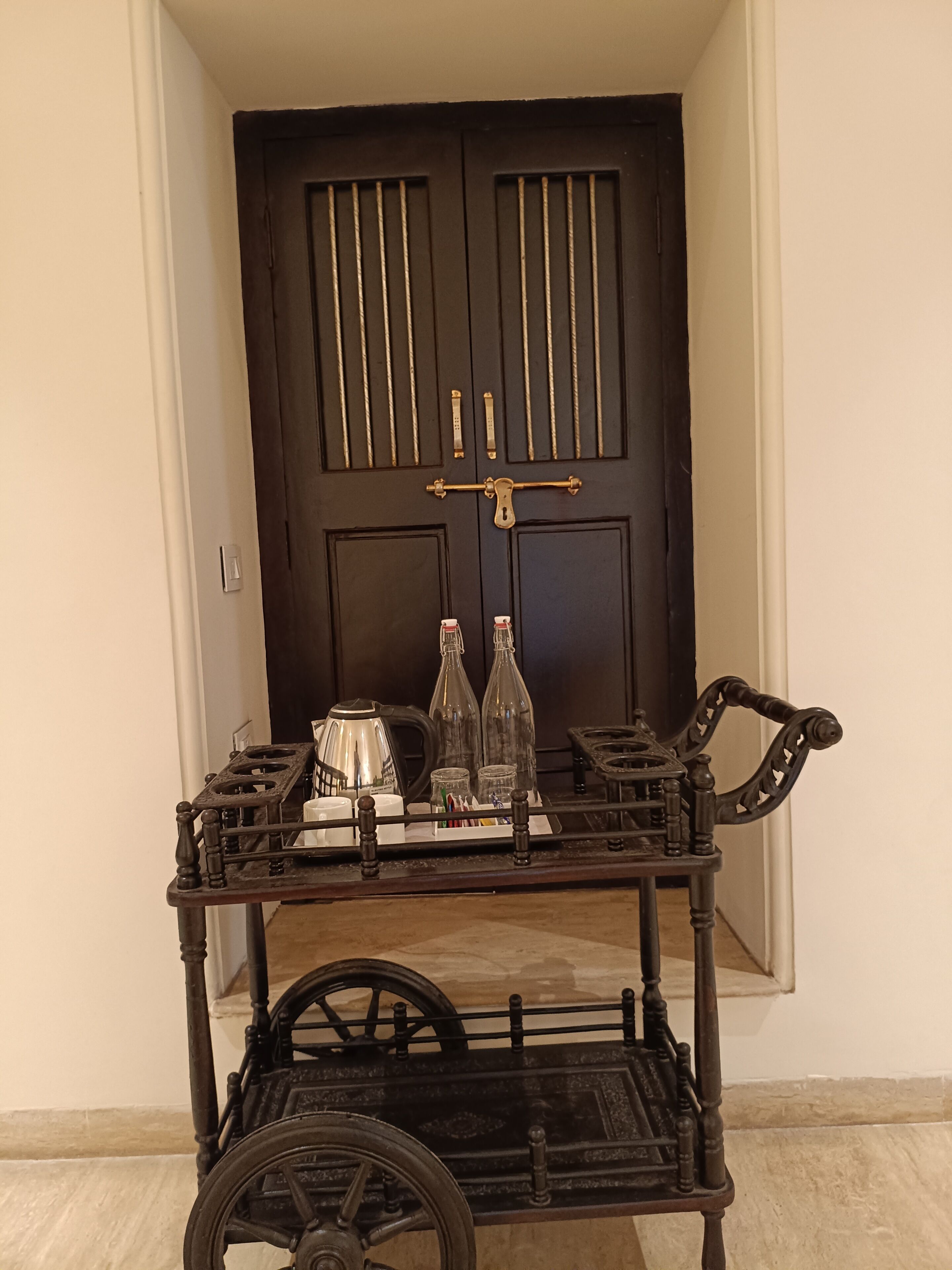 Una puerta de madera oscura con paneles verticales y un tirador de latón. Frente a la puerta, un carro metálico negro sobre ruedas que contiene un hervidor, dos botellas de vidrio y una pequeña mesa con una tetera y tazas.
