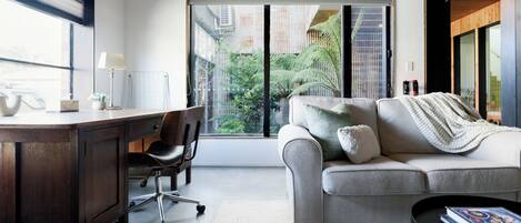 A modern living room with a desk, a comfortable sofa, and a view of a garden through large windows.