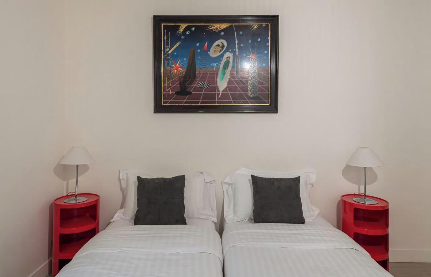 Ein Hotelzimmer mit zwei Betten, jedes mit einem Kopfkissen, sowie ein roter Nachttisch mit einer Lampe. Über den Betten hängt ein gerahmtes Kunstwerk.