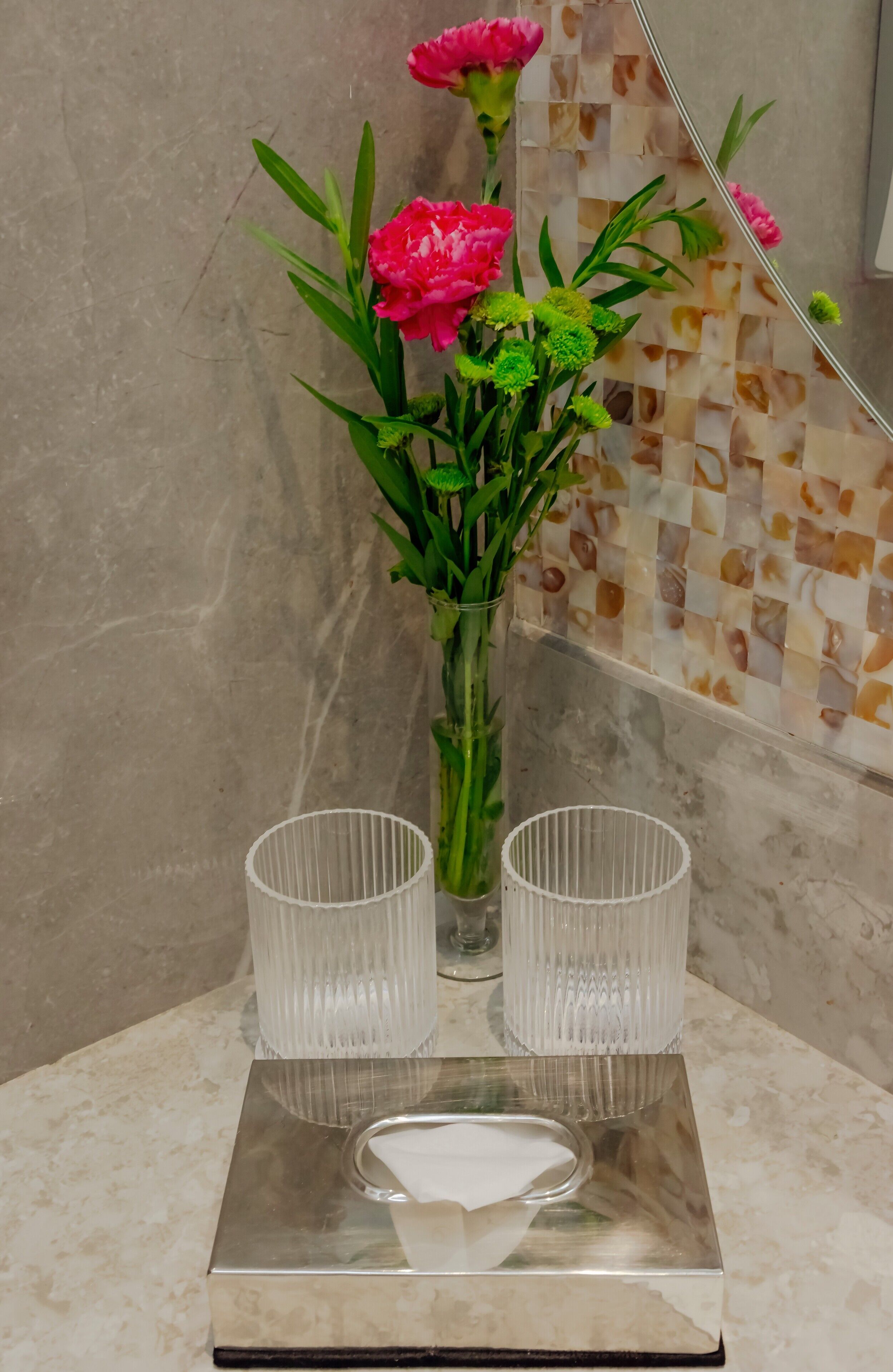 A bathroom with a marble countertop, a glass vase with pink flowers, and two clear glass cups.