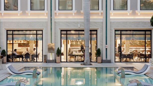 An indoor swimming pool with lounge chairs, a palm tree, and a restaurant with glass doors. - The Tivoli New Delhi (New Delhi)