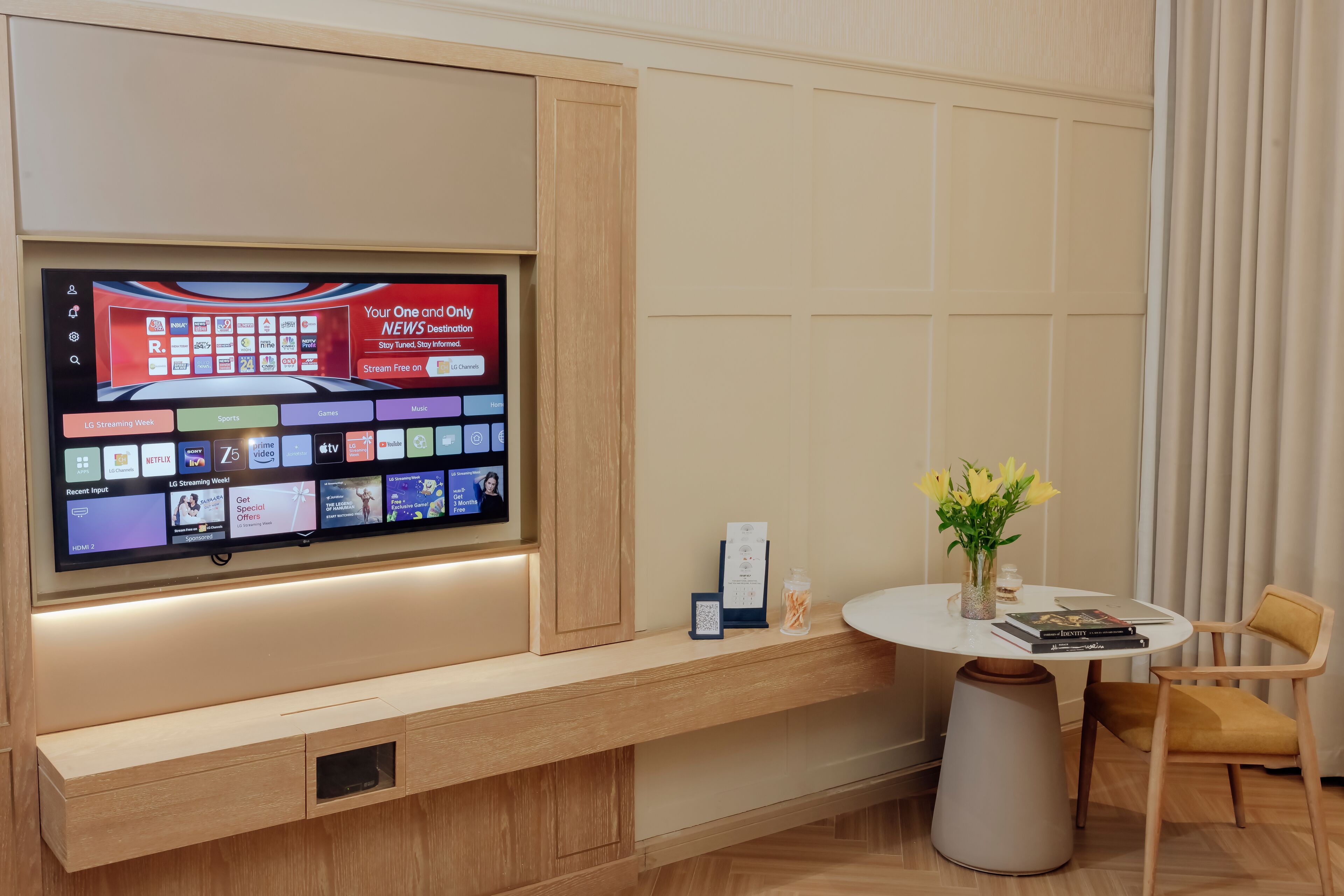 A hotel room with a large flat-screen TV mounted on the wall displaying various streaming services. There is a small table with a vase of flowers, a chair, and some books.