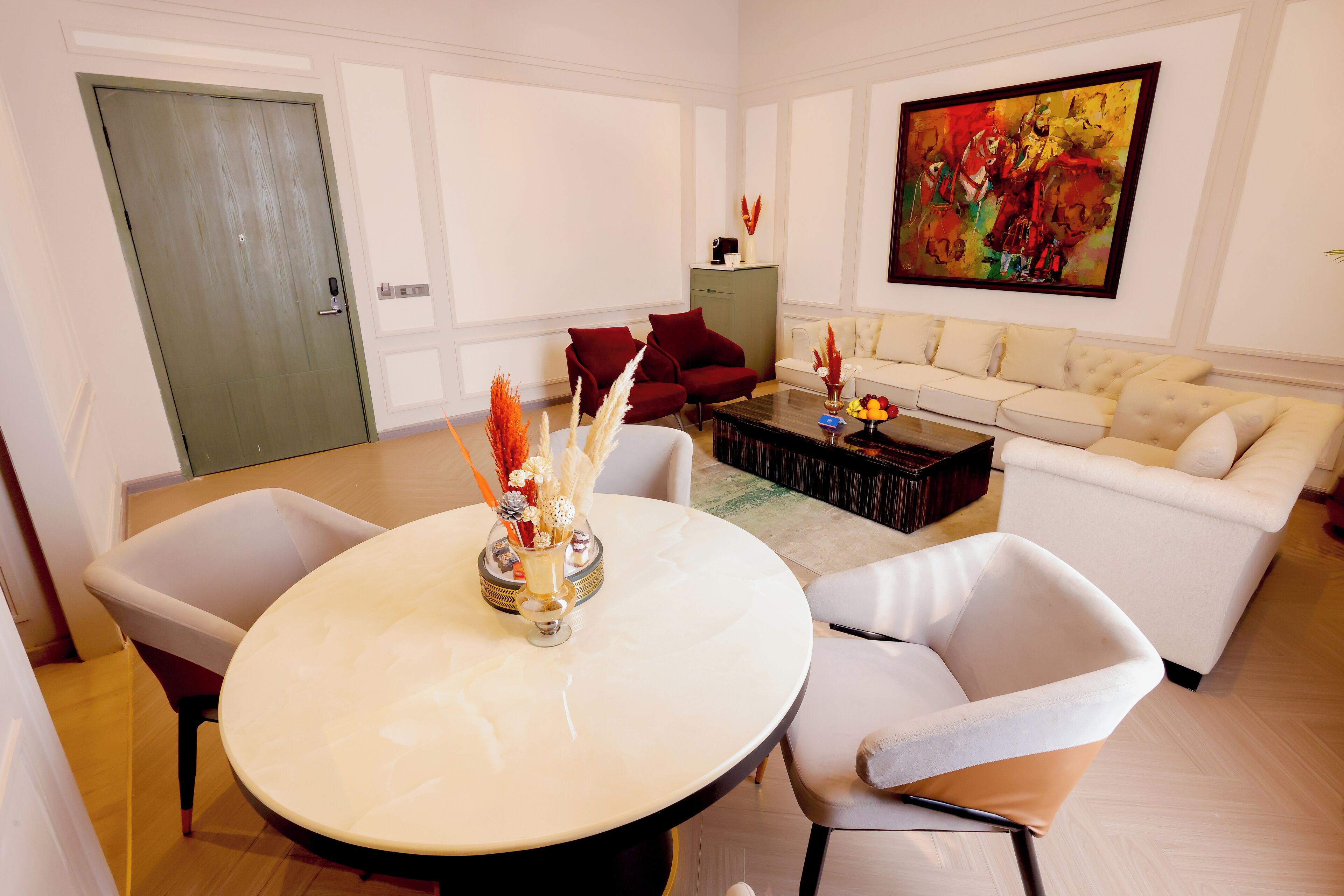 A modern living room with a round dining table, two white chairs, a red sofa, and a large abstract painting on the wall.