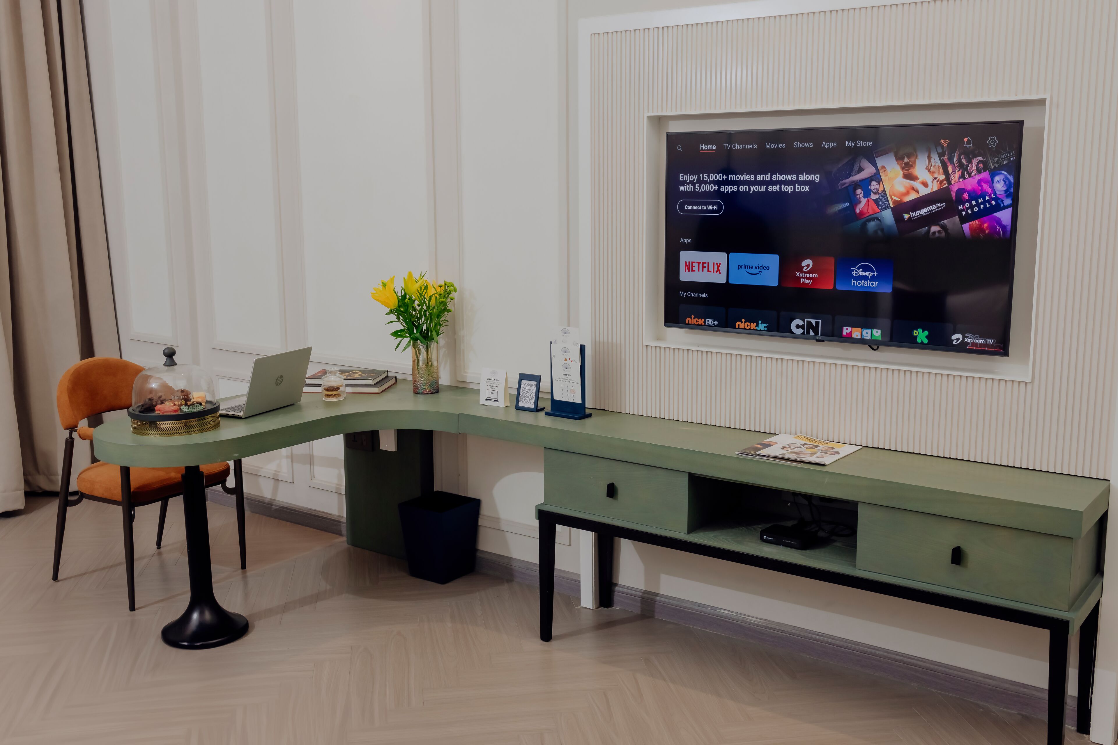 Une chambre d’hôtel moderne avec un bureau incurvé, un ordinateur portable, un vase de fleurs et un téléviseur à écran plat fixé au mur, diffusant des services de streaming.