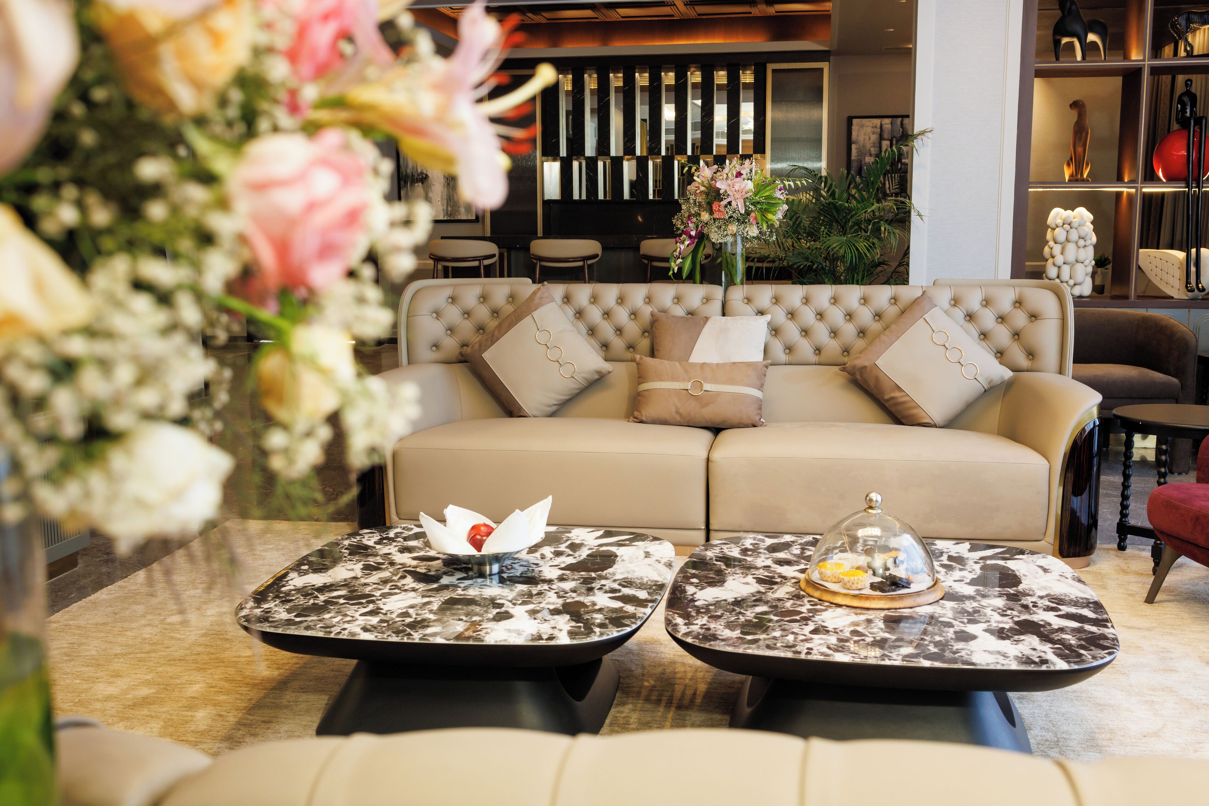 A modern living room with a beige sofa, two square coffee tables with marble tops, and a floral arrangement in the foreground.