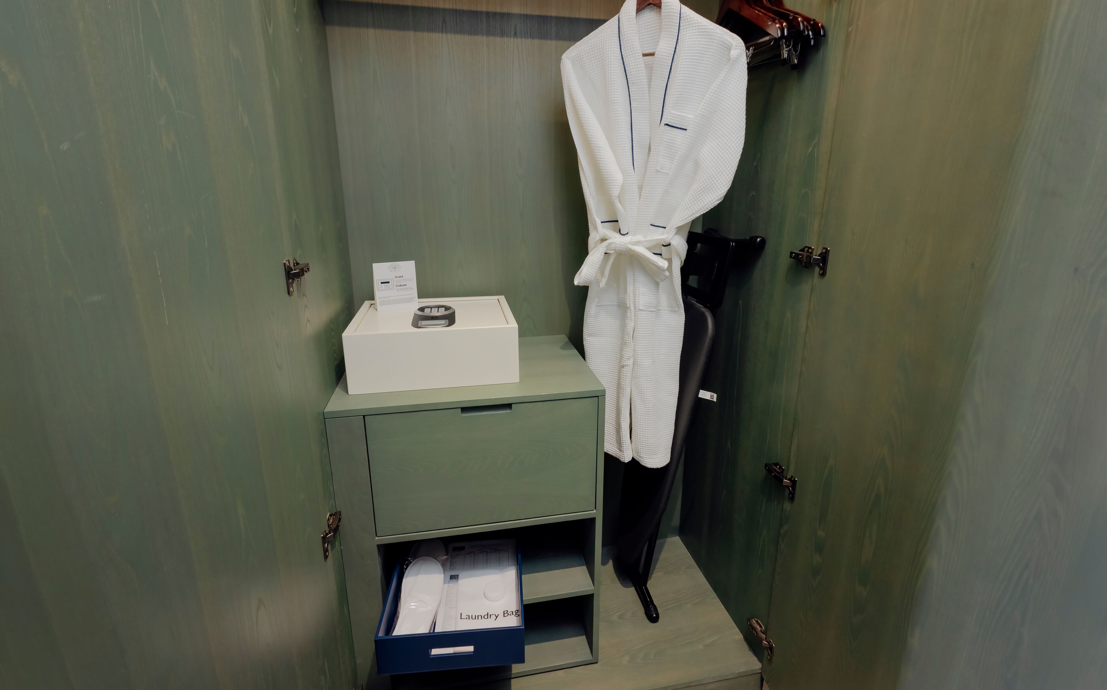 A bathroom with a green cabinet, a white robe hanging on the wall, a black chair, and a blue laundry bag drawer.