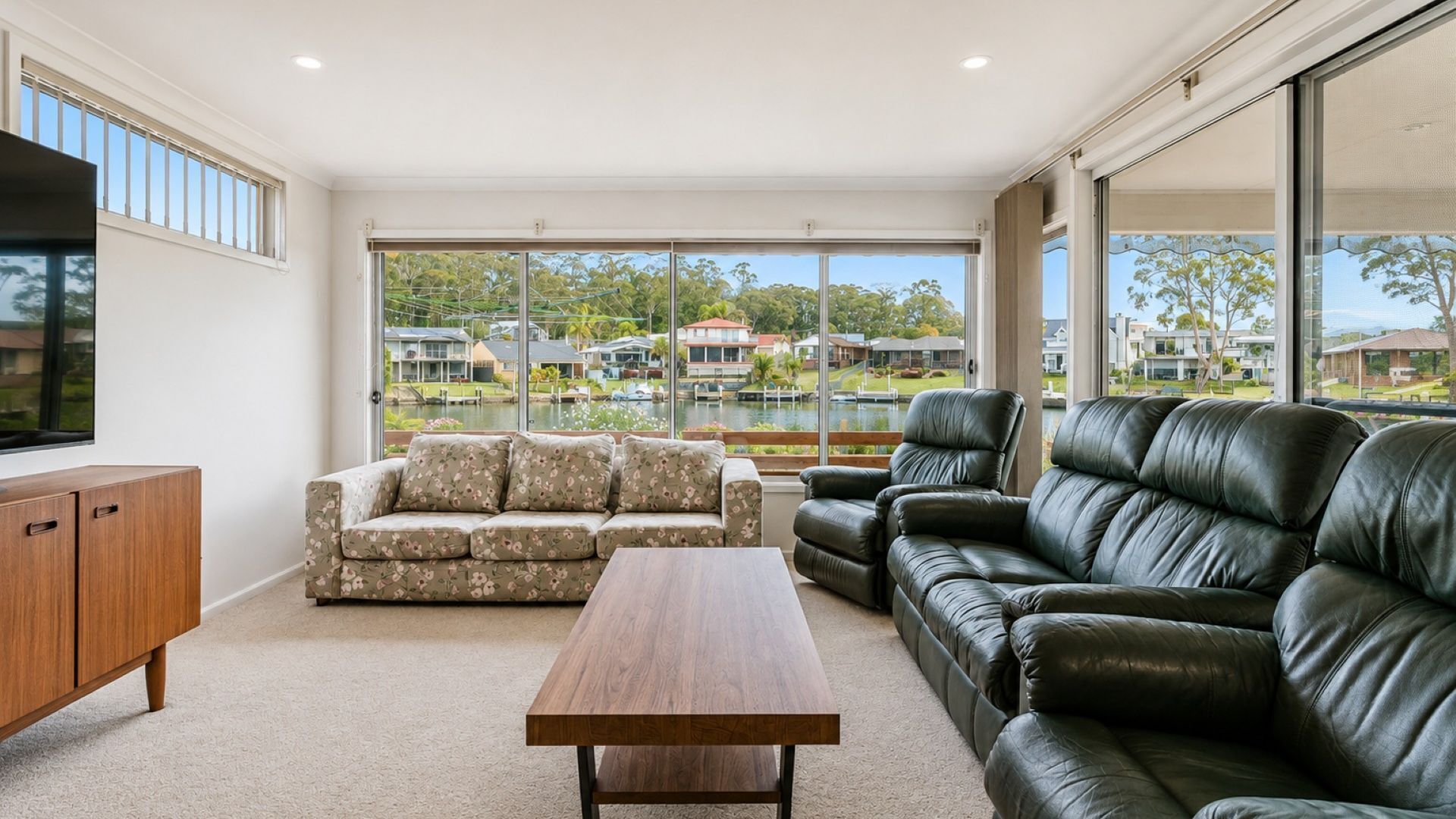 A spacious living room with a wooden coffee table, a floral sofa, and a black leather couch. Large windows offer a view of a residential area.