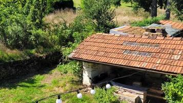 Une maison de campagne en pierre, avec un toit de tuiles, entourée d’une végétation luxuriante et sous un ciel bleu azur.