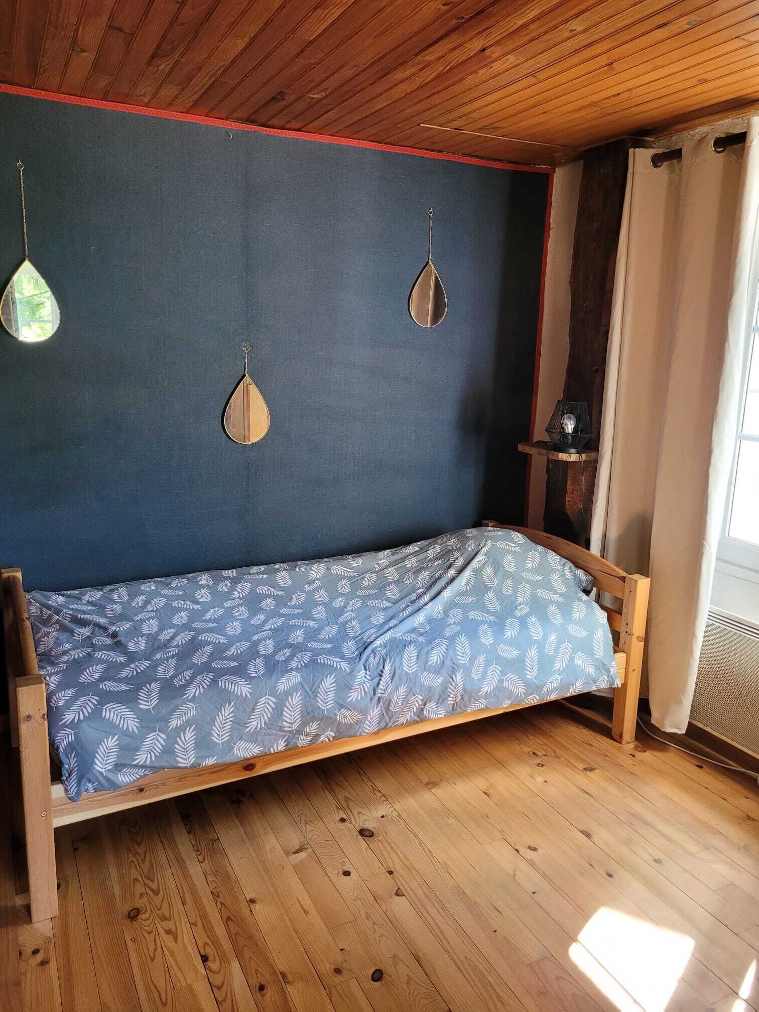 Una habitación pequeña con cama individual, suelo de madera, pared azul y ventana con cortinas.