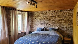 A bedroom with a stone wall, a wooden ceiling, a bed with a blue patterned bedspread, and a window with curtains.