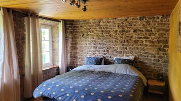 Une chambre avec un mur en pierre, un plafond en bois, un lit recouvert d’une courtepointe à motifs bleus et une fenêtre avec des rideaux.