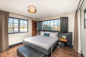Quarto moderno com cama grande, um banquinho, mesa de cabeceira e vista para o exterior.