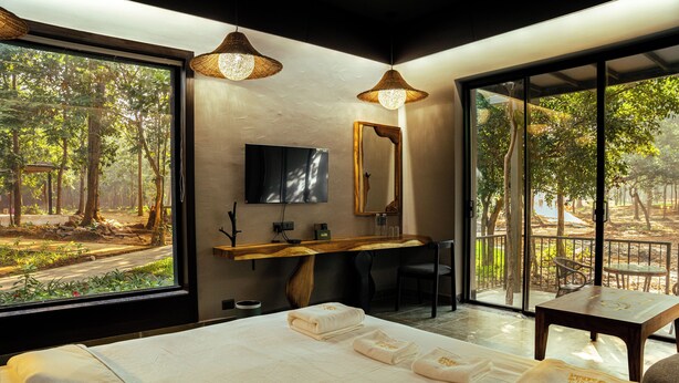 Habitación de hotel con una cama grande, un escritorio, una silla y vistas a un bosque a través de amplias ventanas.