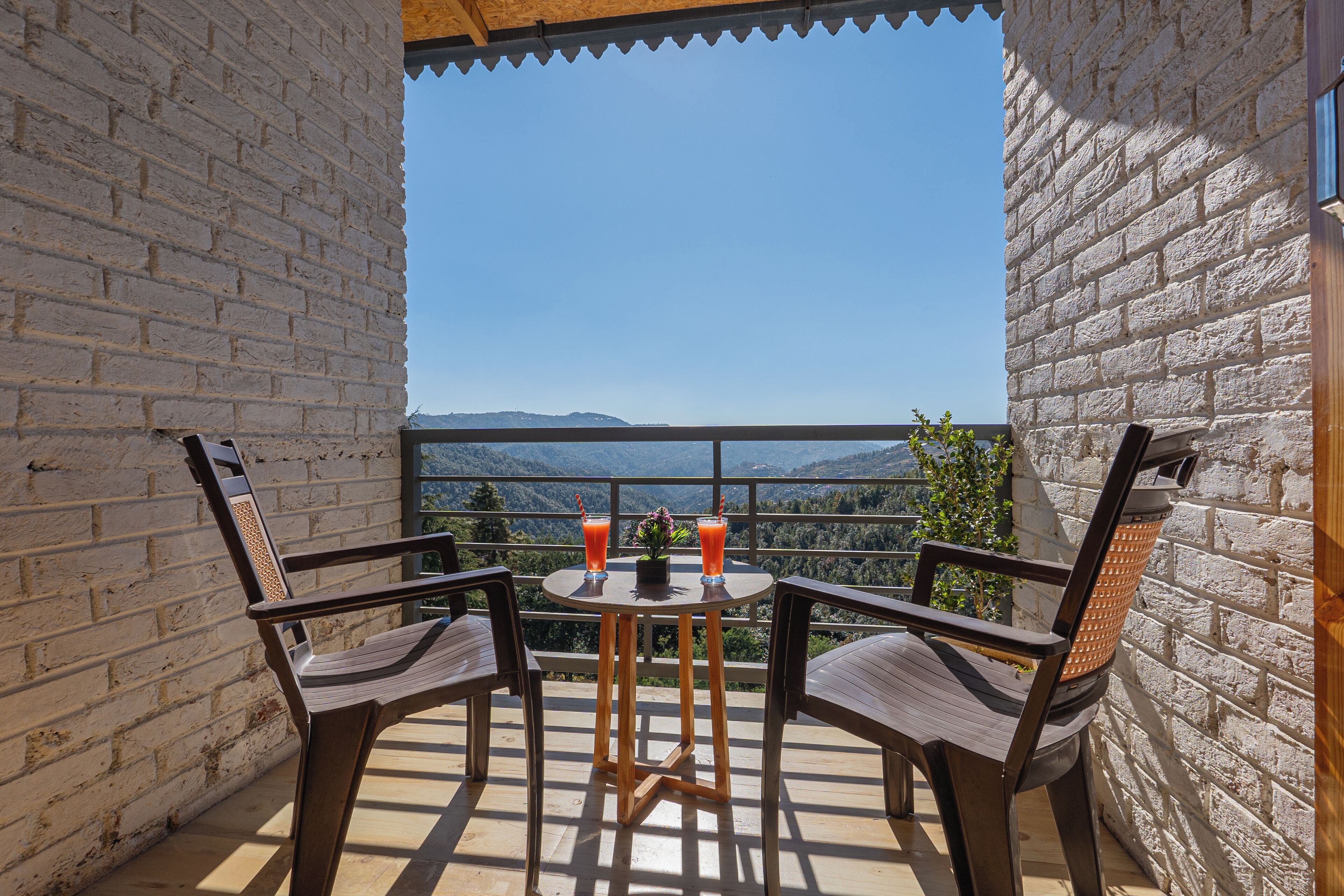 A balcony with two chairs, a small table, and two drinks. The balcony overlooks a mountainous landscape.