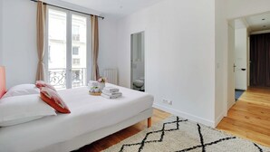 Un dormitorio con cama, ventana con cortinas, espejo y piso de madera.