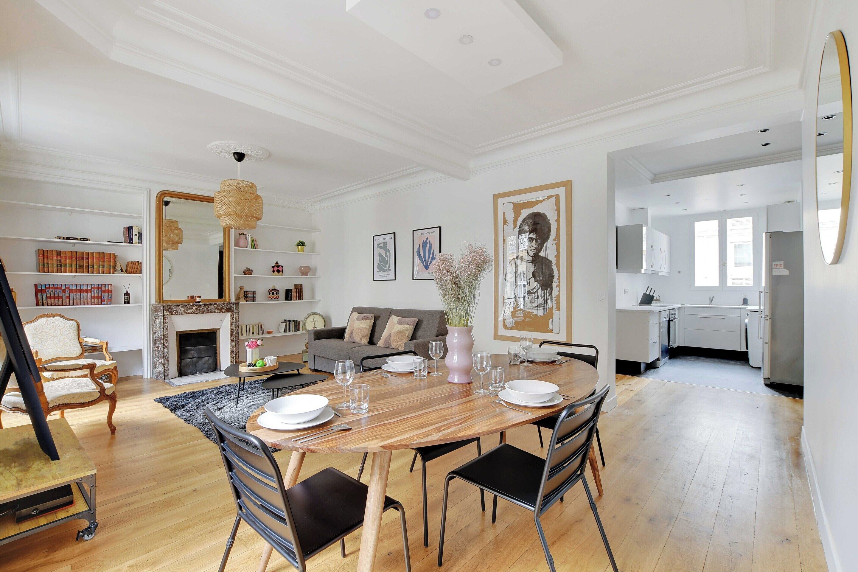 A spacious living room with a wooden dining table, black chairs, a fireplace, and a kitchen in the background.