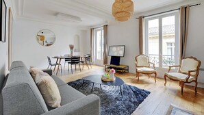 Una sala de estar con un sofá gris, una mesa redonda y dos sillones. Hay un comedor con una mesa y sillas, y un televisor sobre un mueble.
