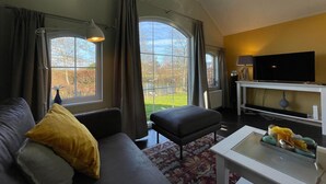 A living room with a sofa, ottoman, TV, and a window with curtains. - Tranquil Lakeside Stay for 4 Guests (Sint Nicolaasga)