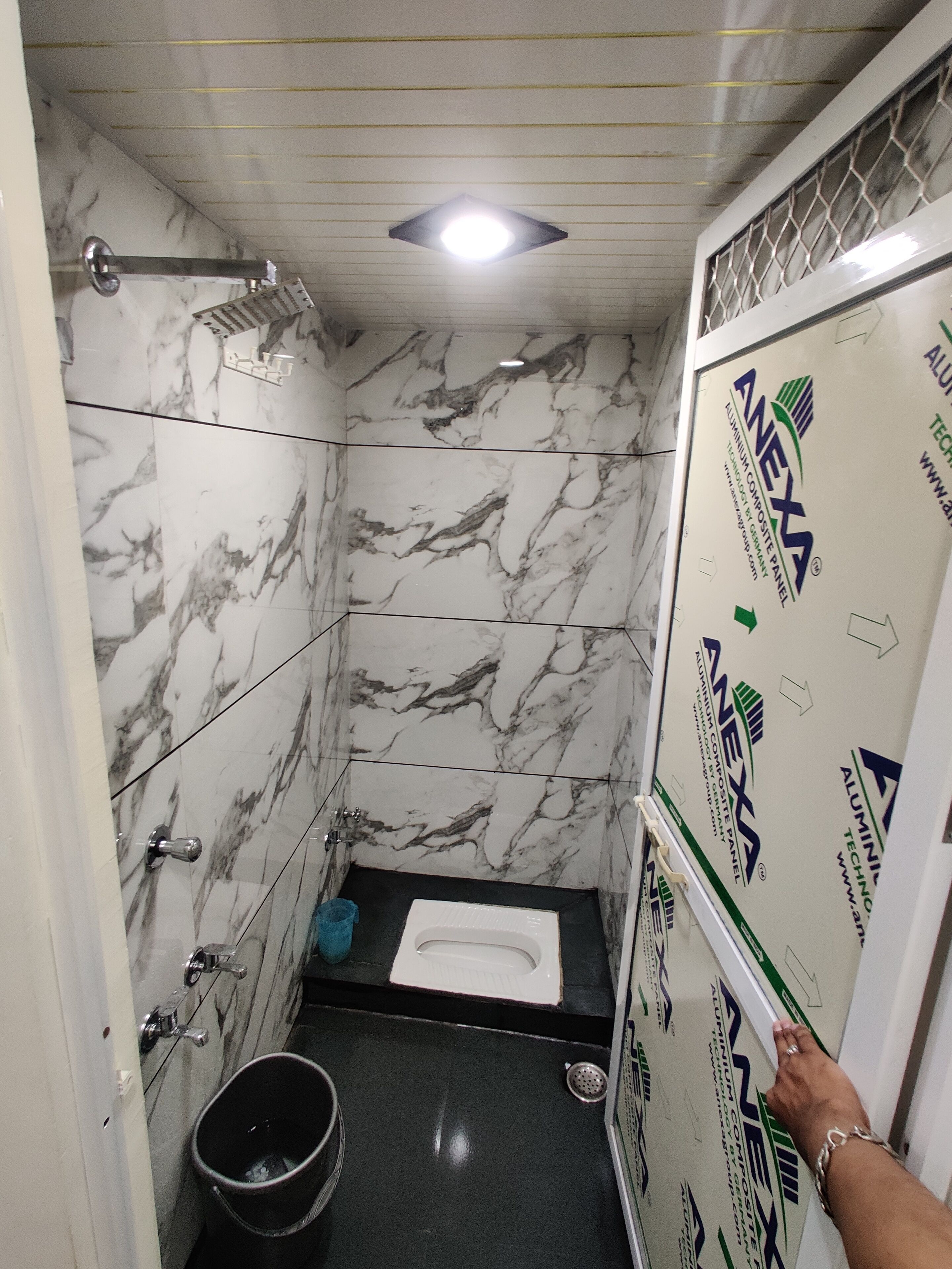 A bathroom with a toilet, a shower, and a sink. The walls are covered in white marble tiles with gray veining. There is a blue trash bin next to the toilet. The ceiling has a recessed light fixture.