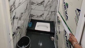 Une salle de bain avec des toilettes, une douche et un lavabo. Les murs sont recouverts de carreaux de marbre blanc à veines grises. Une poubelle bleue se trouve à côté des toilettes. Le plafond est doté d’un luminaire encastré.