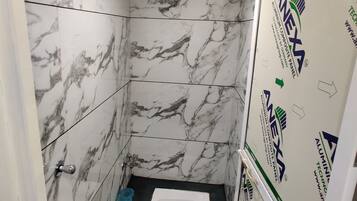 A bathroom with a toilet, a shower, and a sink. The walls are covered in white marble tiles with gray veining. There is a blue trash bin next to the toilet. The ceiling has a recessed light fixture.