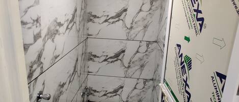 A bathroom with a toilet, a shower, and a sink. The walls are covered in white marble tiles with gray veining. There is a blue trash bin next to the toilet. The ceiling has a recessed light fixture.