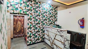 Un espace d’accueil avec un mur orné d’un motif géométrique, un comptoir de réception en marbre, un extincteur et une porte décorée de motifs végétaux.